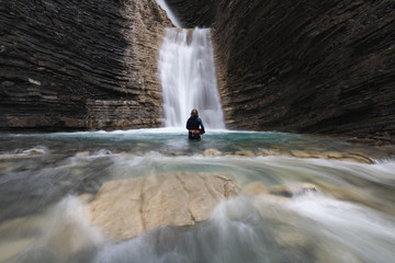 Woman in a canyoning costume in the turquoise water submerged to the waist of a waterfall in a mountain ravine with river rocks in the foreground. Long exposition
