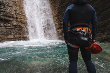 lonely woman with canyoning costume and a red helmet in her hand in the foreground inside the turquoise water in a waterfall of a mountain canyon