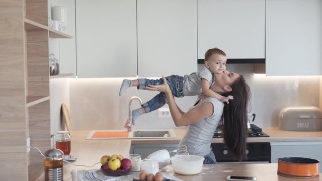 Young beautiful mother and her son are standing in a kitchen and playing together. Woman is holding her child on a hands and tossing him.