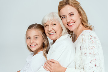 smiling granddaughter, mother and grandmother hugging isolated on grey
