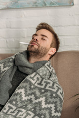 sick man, wrapped in blanket, sitting on sofa with closed eyes and measuring temperature