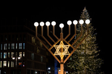 Huge illuminated menorah with round lamps and lighted Jewish symbol David's star on blurry background of Christmas tree in Berlin Germany. Night cityscape with Hanukah decorations