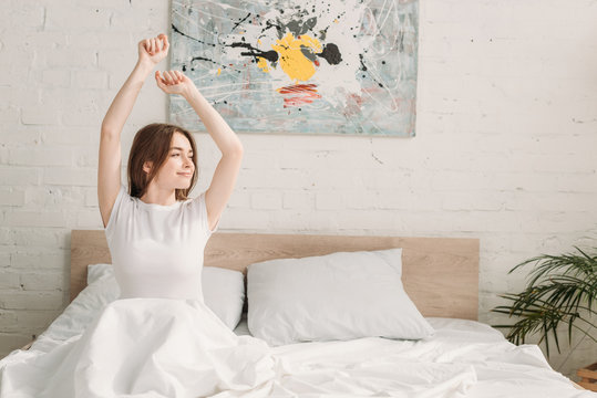 Happy Girl Looking Away While Sitting In Bed And Stretching In Morning