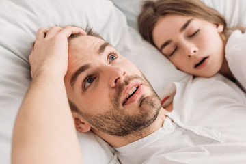 awakened man holding hand on head while lying in bed near snoring wife