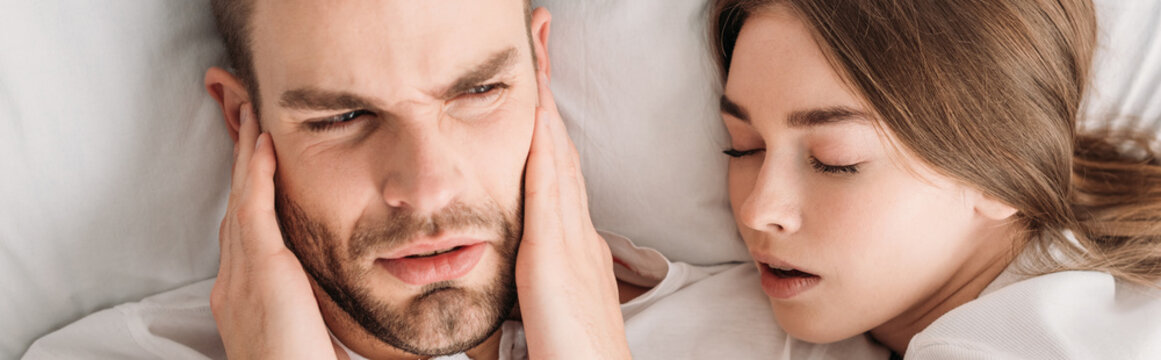 top view of exhausted man plugging ears with hands while lying in bed near snoring wife, panoramic shot