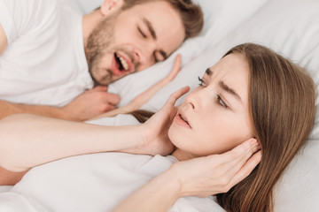 selective focus of woman plugging ears with hands while lying in bed near snoring husband
