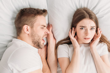 top view of dissatisfied woman plugging ears with hands and looking at camera while lying in bed...
