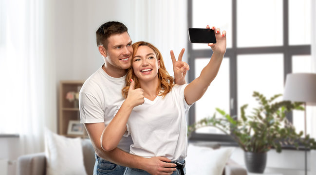 relationships, technology and people concept - happy couple in white t-shirts taking selfie smartphone and showing peace by over home background
