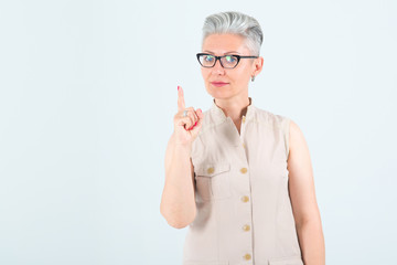 Middle aged woman in glasses, points a finger up, standing on a light background.