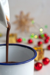 Close-up of jug serving chocolate over white cup with red Christmas decorations on bokeh, on white wooden background in verical