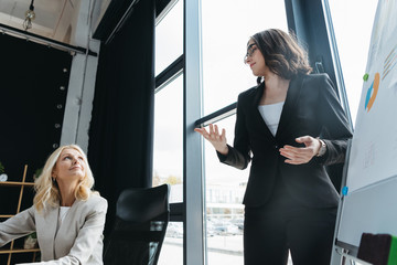 young manager talking to mature businesswoman while standing near flipchart and showing explain gesture