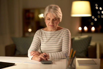 technology, old age and people concept - senior woman with laptop counting on calculator and filling tax form at home in evening