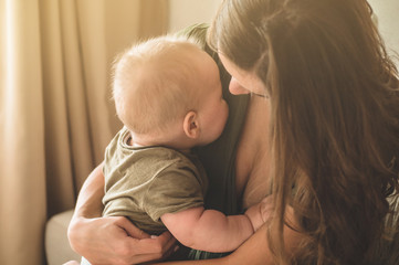 Home portrait of a baby boy with mother on the bed. Mom holding and kissing her child. 