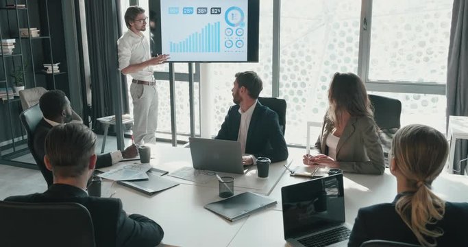 Young Attractive Bearded Man Explaining his Ideas to Teamwork in the Office by the Interactive Board. Colleagues listening Attentively Manager. Noting on Papers Important Ideas. Busy Business People.