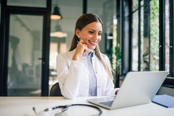 Portrait of a smiling female doctor looking at laptop in medical office.
