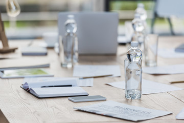 selective focus of bottle with water near smartphone, notebook and documents on wooden table in meeting room