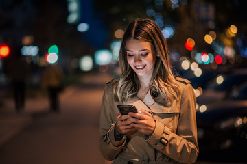Portrait of a smiling young woman using smart phone at night. - obrazy, fototapety, plakaty
