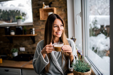 Beautiful smiling brunette looking through window and drinking coffee in the kitchen. - Powered by Adobe