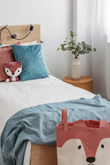 Green eucalyptus in stylish vase on wooden nightstand next to single bed with toy, and blue and red bedding