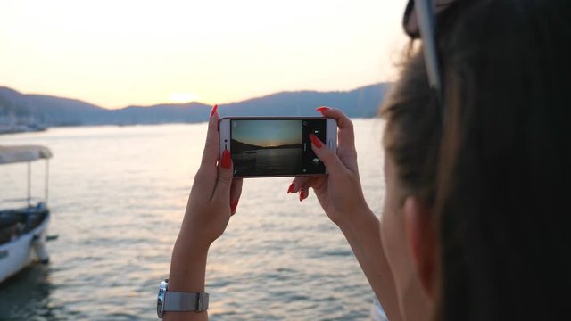 Woman making photo of scenic marine landscape with golden sunset on his smartphone. Girl using phone to get beautiful pictures of seascape. Lady enjoying vacation resting on seashore at evening time