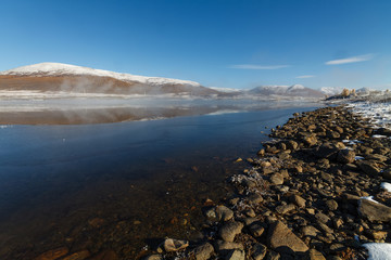 Obraz premium Mountain lake in Siberia in September with pebbles on the shore and reflection of snowy hills and blue sky