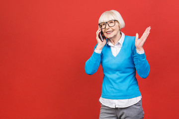 Fototapeta premium Phone conversation. Positive senior aged woman smiling while talking on the phone isolated over red background.