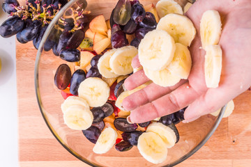 Cooking a vegetarian healthy fruit salad - female hand puts sliced bananas in a glass bowl with berries and fruits