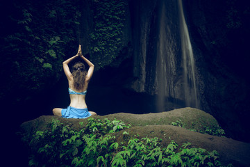 Woman sitting on the rock, practicing yoga. Young woman raising arms with namaste mudra near waterfall. Leke Leke waterfall, Bali. View from back.