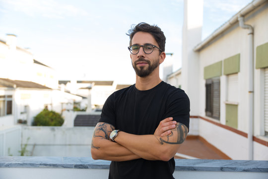 Confident stylish guy with tattoos posing on apartment balcony or terrace. Young man in glasses standing outside with arms crossed and looking at camera. Male portrait concept