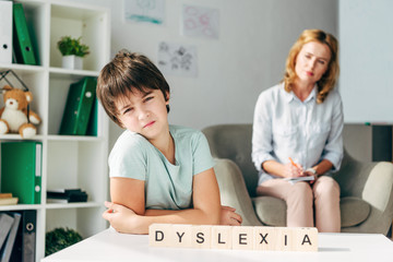 selective focus of sad kid with dyslexia sitting at table with wooden cubes with lettering dyslexia