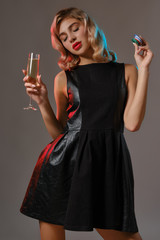 Blonde girl in black dress holding glass of champagne and chips, posing against gray background. Gambling, poker, casino. Close-up.