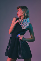 Blonde girl in black stylish dress holding some money and chips, posing against colorful background. Gambling entertainment, poker, casino. Close-up.
