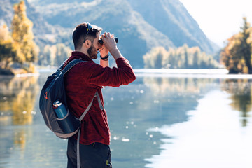 Hiker young man traveler with backpack looking the landscape with binoculars in the lake.