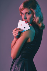 Blonde girl in black stylish dress showing two playing cards, posing against colorful background. Gambling entertainment, poker, casino. Close-up.