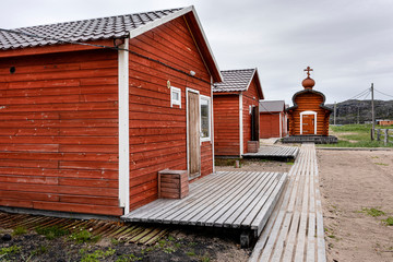 Russia, Arctic, Kola Peninsula, Barents Sea, Teriberka: New red wooden houses in the center of the old Russian settlement small fishing village with small chapel, boardwalk and grey cloudy sky.