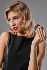 Blonde girl in black leather dress holding some colorful chips, posing against gray background. Gambling entertainment, poker, casino. Close-up.