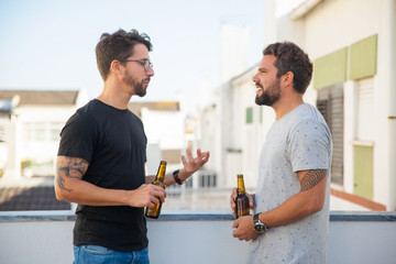 Two male friends excited with discussion over bottle of beer on outdoor terrace. Two young men in...