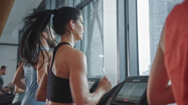 Beautiful Athletic Sports Woman Wearing Wireless Headphones, Listens to a Podcast or Sport Music Playlist while Running on a Treadmill. Fit Athletes Training in the Gym. Slow Motion Side View