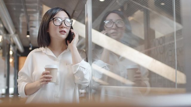 Medium shot of young pretty Asian businesswoman wearing office clothes standing leaning on glass wall, holding paper coffee cup while talking on phone