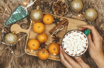 Wooden board with tangerines and cup of cocoa with woman hands on it, golden balls, cones, cinnamon and anice star, caramel on a background of brown fur. New Year flat lay