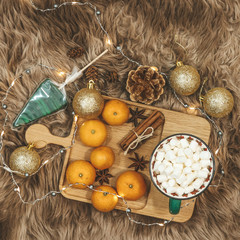 Fototapeta na wymiar Wooden board with tangerines and cocoa with marshmallows, golden balls, cones, cinnamon and anice star, caramel on a background of brown fur. New Year flat lay