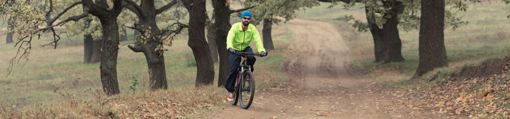 Fototapeta na wymiar A guy rides a bicycle in an oak forest on a dirt road. Biker in a light green jacket on a modern mountain carbon bike.