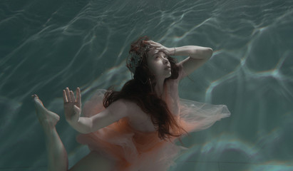 A girl with long dark hair swims underwater in a pink dress and with a crown on her head, like an...