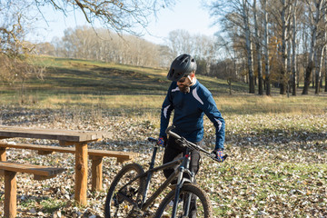 Fototapeta premium Cyclist in pants and fleece jacket on a modern carbon hardtail bike with an air suspension fork rides off-road. The guy is resting on a bench in the autumn park.