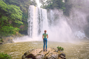 Young blond woman standing on a rock watching the landscape with a waterfall in the middle of the jungle