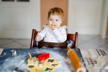 Cute little baby girl baking gingerbread Christmas cookies at home. Adorable blond happy healthy child having fun in domestic kitchen. Traditional leisure with kids on Xmas. Toddler tasting dough.