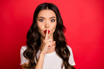Photo of curly wavy brown haired woman in white t-shirt showing you shh sign to stop your talking with forefinger touching lips isolated vivid color background