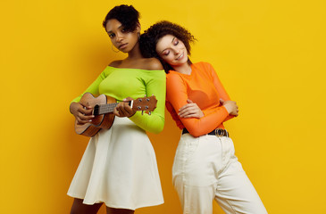 Two young beautiful hipster girls in colorful summer clothes with small guitar