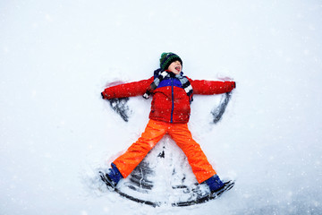 Cute little kid boy in colorful winter clothes making snow angel, laying down on snow. Active outdoors leisure with children in winter. Happy healthy child having fun and laughing outdoors