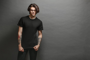 Hipster handsome male model with glasses wearing black blank t-shirt and black jeans with space for your logo or design in casual urban style - obrazy, fototapety, plakaty